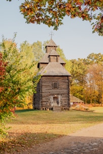 A rustic wooden building with a steep, gabled roof stands in the middle of a park surrounded by trees in their autumn colors. The building seems to be constructed from logs, giving it a quaint and historical appearance. Bright fall leaves are scattered on the ground and hang from branches above, framing the structure.
