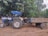 A blue and white tractor with a canopy is connected to a rustic trailer. The trailer has decorative artwork and text in a local script on its side. The scene is set outdoors with a background of lush green trees and a dirt ground.