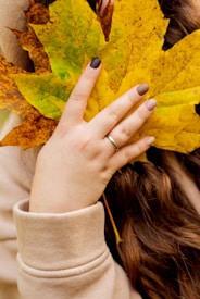 A close-up of a hand with painted nails holding a large yellow maple leaf. The person is wearing a beige sweatshirt and has long, wavy brown hair.