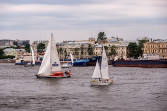 The image depicts a waterfront scene with numerous sailboats in the water. There are buildings in the background, including both modern and classical architecture. The sky is overcast, and the water appears to be calm. Several people can be seen on the boats, indicating recreational sailing activity.