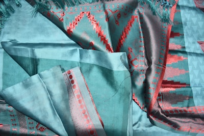 A fabric with intricate patterns featuring vibrant red geometric designs on a soft teal background. The material appears silky with a sheen, and various folds suggest a textured, rich textile.