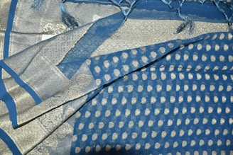 An intricately patterned fabric prominently featuring a mix of blue and silver colors. The design includes small circular motifs and elaborate borders with geometric and decorative patterns. The fabric has a shiny texture, and fringes are visible at one edge.