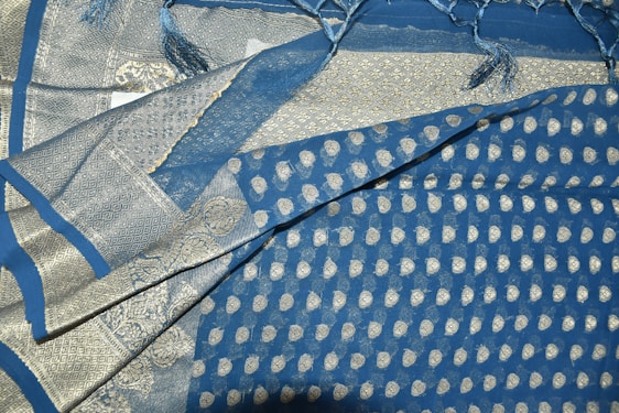 An intricately patterned fabric prominently featuring a mix of blue and silver colors. The design includes small circular motifs and elaborate borders with geometric and decorative patterns. The fabric has a shiny texture, and fringes are visible at one edge.