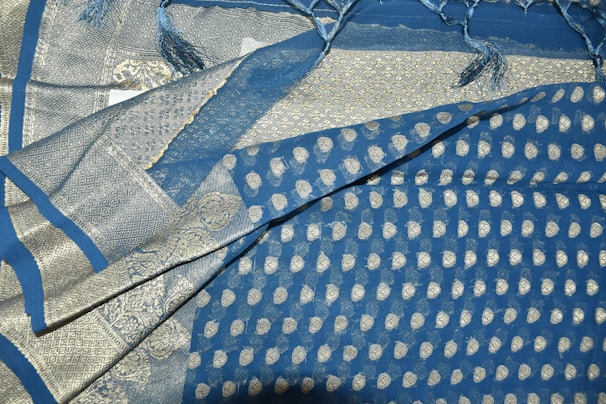 An intricately patterned fabric prominently featuring a mix of blue and silver colors. The design includes small circular motifs and elaborate borders with geometric and decorative patterns. The fabric has a shiny texture, and fringes are visible at one edge.