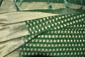 A richly textured fabric with ornate gold patterns and motifs on a deep green background. The design features intricate paisleys and geometric shapes, bordered by a gilded edge with detailed stitching. Fringes adorn the fabric's edge, adding an elegant touch.
