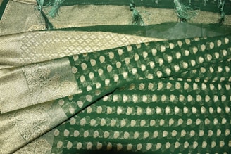 A richly textured fabric with ornate gold patterns and motifs on a deep green background. The design features intricate paisleys and geometric shapes, bordered by a gilded edge with detailed stitching. Fringes adorn the fabric's edge, adding an elegant touch.