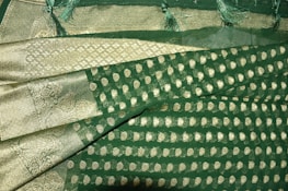 A richly textured fabric with ornate gold patterns and motifs on a deep green background. The design features intricate paisleys and geometric shapes, bordered by a gilded edge with detailed stitching. Fringes adorn the fabric's edge, adding an elegant touch.
