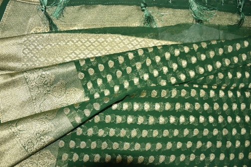 A richly textured fabric with ornate gold patterns and motifs on a deep green background. The design features intricate paisleys and geometric shapes, bordered by a gilded edge with detailed stitching. Fringes adorn the fabric's edge, adding an elegant touch.