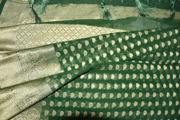 A richly textured fabric with ornate gold patterns and motifs on a deep green background. The design features intricate paisleys and geometric shapes, bordered by a gilded edge with detailed stitching. Fringes adorn the fabric's edge, adding an elegant touch.