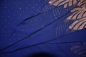 A fabric with a deep blue background is adorned with intricate gold patterns and small sparkling dots. The patterns resemble fireworks or radial designs, creating an elegant and luxurious look.