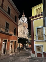 A nighttime scene featuring a cobblestone street flanked by colorful colonial-style buildings. In the background, an illuminated historic church tower stands prominently, showcasing intricate architectural details. The street is quiet and appears empty of people, with a few lit storefronts visible.