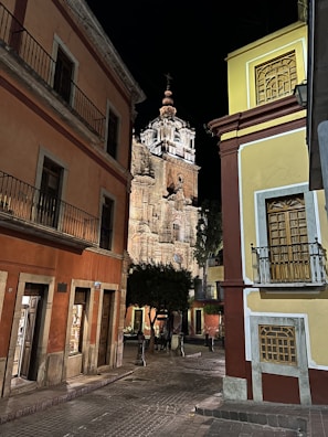 A nighttime scene featuring a cobblestone street flanked by colorful colonial-style buildings. In the background, an illuminated historic church tower stands prominently, showcasing intricate architectural details. The street is quiet and appears empty of people, with a few lit storefronts visible.