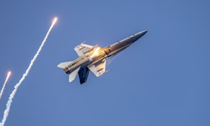 A fighter jet emits bright flares against a clear blue sky, with dramatic lighting highlighting the aircraft's sleek design.