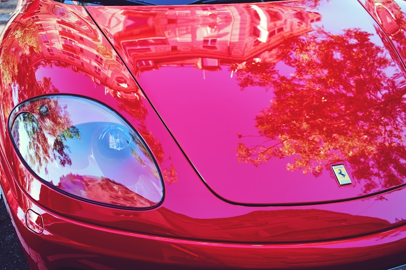 A close-up view of a highly polished red sports car with reflections of trees and buildings visible in the glossy finish. The image showcases the sleek design of the car and its shiny surface, including a headlight and an emblem prominently displayed on the hood.
