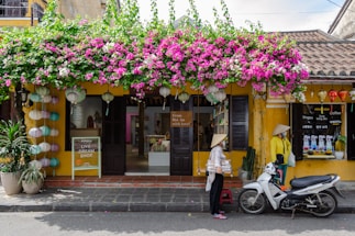 A quaint shop with a vibrant display of pink and white flowers hanging above its entrance, complemented by colorful lanterns. The building features an inviting yellow facade with dark wooden shutters and a tiled roof. Two people wearing traditional hats are standing nearby, one of them beside a white scooter parked on the road.
