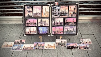 A collection of colorful postcards featuring famous landmarks, likely from a cityscape, displayed on a metal stand on a pavement. The images include various scenes with notable structures, each postcard showcasing artistic views with vibrant skies and architectural details. A small sign indicates a price of £15.