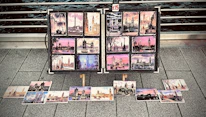 A collection of colorful postcards featuring famous landmarks, likely from a cityscape, displayed on a metal stand on a pavement. The images include various scenes with notable structures, each postcard showcasing artistic views with vibrant skies and architectural details. A small sign indicates a price of £15.