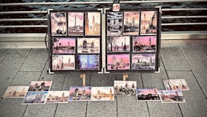A collection of colorful postcards featuring famous landmarks, likely from a cityscape, displayed on a metal stand on a pavement. The images include various scenes with notable structures, each postcard showcasing artistic views with vibrant skies and architectural details. A small sign indicates a price of &pound;15.