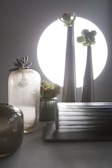 A composition of sleek, metallic and glass vases with succulents arranged inside, set against a large, bright circular light source in the background. The arrangement includes a variety of textures from smooth to frosted surfaces, contributing to a modern and minimalistic aesthetic.