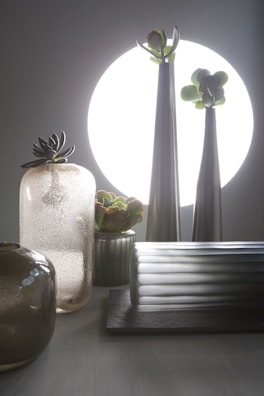 A composition of sleek, metallic and glass vases with succulents arranged inside, set against a large, bright circular light source in the background. The arrangement includes a variety of textures from smooth to frosted surfaces, contributing to a modern and minimalistic aesthetic.