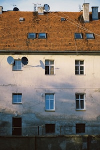 A building facade with a weathered look, featuring multiple windows with both double and single panes. There are several satellite dishes affixed to the exterior. The roof is covered with brown tiles and has chimneys and small attic windows. The walls exhibit signs of wear, with peeling paint and visible cracks.