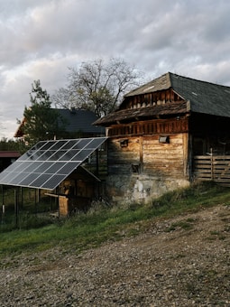 A rustic wooden barn with a stone base is situated on a grassy hill. Two solar panels are installed nearby, supported by a metal framework. In the background, there are trees with sparse foliage and another building partially visible. The sky is overcast with clouds.