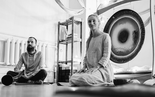 Two individuals are sitting on the floor in a room that appears to be used for meditation or relaxation. The woman is smiling and sitting in front of a large gong. The man is sitting cross-legged with a calm expression and has a long wooden instrument resting beside him. Shelves in the background hold yoga blocks and books.