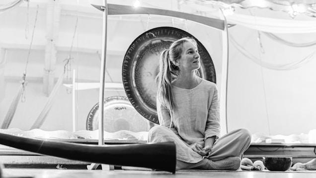 A person with long hair sits cross-legged on the floor, surrounded by musical instruments like a large gong and a didgeridoo. The scene has an artistic and relaxed atmosphere, with soft lighting and a blend of natural elements.