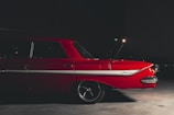 A vintage red car is parked indoors with dim lighting. The vehicle has a white stripe along the side and features classic design elements, including a prominent tail fin. The word 'Impala' is visible on the side of the car.