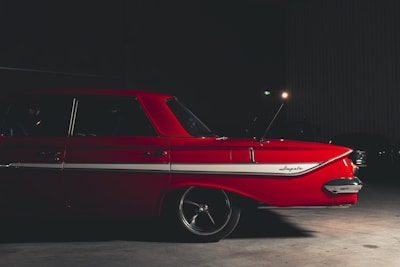 A vintage red car is parked indoors with dim lighting. The vehicle has a white stripe along the side and features classic design elements, including a prominent tail fin. The word 'Impala' is visible on the side of the car.