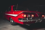 A classic red car with a sleek design is parked in a dimly lit environment. Its rear lights are illuminated, casting a subtle glow. The car features chrome details and a distinctive stripe along the side. The branding reads 'Impala' on the side, denoting a vintage model.