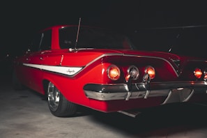 A classic red car with a sleek design is parked in a dimly lit environment. Its rear lights are illuminated, casting a subtle glow. The car features chrome details and a distinctive stripe along the side. The branding reads 'Impala' on the side, denoting a vintage model.
