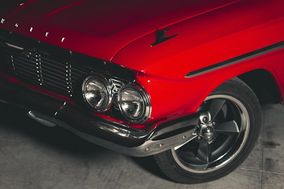 A close-up view of a vintage car highlighting its vibrant red color and chrome details. The image focuses on the car's front section, featuring a prominent grille, round headlights, and a sleek bumper. The wheel is partially visible, showcasing an alloy rim. The car's classic design elements are accentuated by the lighting, creating a sense of nostalgia.