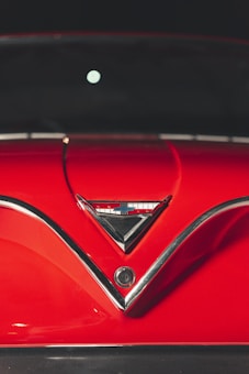 A close-up of a vintage car hood featuring a shiny red surface with chrome trim. At the center of the hood is a triangular emblem with chrome detailing, reflecting sleek design elements typical of classic automobiles.