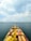 A large cargo ship loaded with colorful containers is navigating through an open sea under a partly cloudy sky. Numerous other ships can be seen scattered in the distance across the water.
