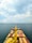A large cargo ship loaded with colorful containers is navigating through an open sea under a partly cloudy sky. Numerous other ships can be seen scattered in the distance across the water.