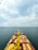 A large cargo ship loaded with colorful containers is navigating through an open sea under a partly cloudy sky. Numerous other ships can be seen scattered in the distance across the water.
