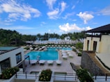 A luxurious resort featuring a large swimming pool surrounded by lounge chairs and umbrellas. Palm trees line the area, with the ocean visible in the background under a partly cloudy sky. Two modern buildings frame the scene, with well-manicured gardens adding to the tropical ambiance.