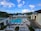 A luxurious resort featuring a large swimming pool surrounded by lounge chairs and umbrellas. Palm trees line the area, with the ocean visible in the background under a partly cloudy sky. Two modern buildings frame the scene, with well-manicured gardens adding to the tropical ambiance.