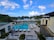 A luxurious resort featuring a large swimming pool surrounded by lounge chairs and umbrellas. Palm trees line the area, with the ocean visible in the background under a partly cloudy sky. Two modern buildings frame the scene, with well-manicured gardens adding to the tropical ambiance.