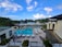 A luxurious resort featuring a large swimming pool surrounded by lounge chairs and umbrellas. Palm trees line the area, with the ocean visible in the background under a partly cloudy sky. Two modern buildings frame the scene, with well-manicured gardens adding to the tropical ambiance.