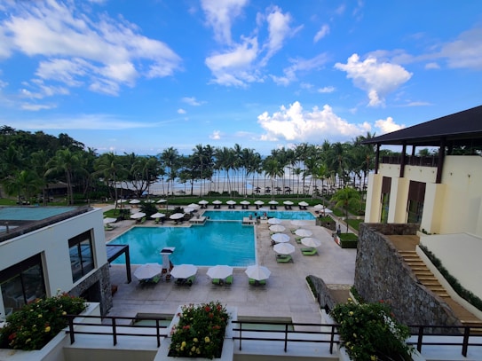A luxurious resort featuring a large swimming pool surrounded by lounge chairs and umbrellas. Palm trees line the area, with the ocean visible in the background under a partly cloudy sky. Two modern buildings frame the scene, with well-manicured gardens adding to the tropical ambiance.