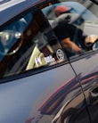 A close-up of a car's side window with stickers attached, including one with the text 'DUBAI DRIVES' and a gear shift pattern. The reflection on the car's surface and the partial view of a person wearing a cap are visible.