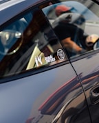 A close-up of a car's side window with stickers attached, including one with the text 'DUBAI DRIVES' and a gear shift pattern. The reflection on the car's surface and the partial view of a person wearing a cap are visible.