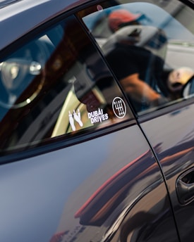 A close-up of a car's side window with stickers attached, including one with the text 'DUBAI DRIVES' and a gear shift pattern. The reflection on the car's surface and the partial view of a person wearing a cap are visible.