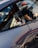 A close-up of a car's side window with stickers attached, including one with the text 'DUBAI DRIVES' and a gear shift pattern. The reflection on the car's surface and the partial view of a person wearing a cap are visible.