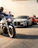 Motorcycles and luxury cars are parked in a line on a street. In the foreground, a person is riding a motorcycle. Behind it, there is a silver sports car followed by a red sports car. Two people are standing next to the red car, talking.