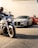 Motorcycles and luxury cars are parked in a line on a street. In the foreground, a person is riding a motorcycle. Behind it, there is a silver sports car followed by a red sports car. Two people are standing next to the red car, talking.