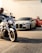 Motorcycles and luxury cars are parked in a line on a street. In the foreground, a person is riding a motorcycle. Behind it, there is a silver sports car followed by a red sports car. Two people are standing next to the red car, talking.