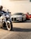 Motorcycles and luxury cars are parked in a line on a street. In the foreground, a person is riding a motorcycle. Behind it, there is a silver sports car followed by a red sports car. Two people are standing next to the red car, talking.